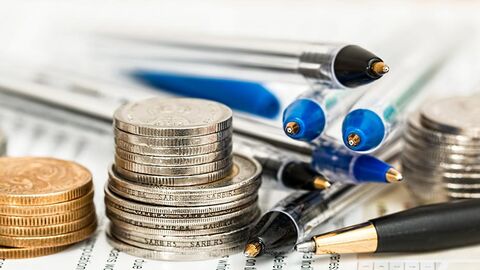 Kleine Münzstapel und verschiedene Kugelschreiber liegen auf einem beschrifteten Papier.