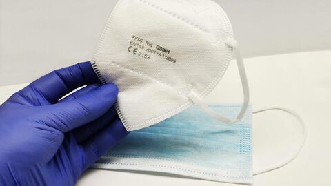 Hand mit blauem Gummihandschuh greift FFP-2-Maske vom Tisch, dort liegt noch eine OP-Maske.