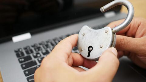 Ein geöffnetes Vorhängeschloss wird von zwei Händen gehalten, im Hintergrund befindet sich ein Laptop.
