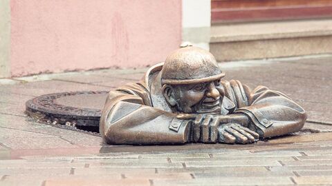 Bronzefigur eines Arbeiters steckt in einem Kanal und lehnt mit beiden Armen auf einer Straße.