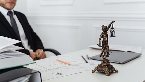 Person sitzt an einem Schreibtisch mit Unterlagen, eine Justitia-Figur steht auf dem Schreibtisch