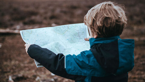 Wegbeschreibung (Symbolbild) Junge steht auf einem Feld und schaut in eine aufgeklappte Landkarte.